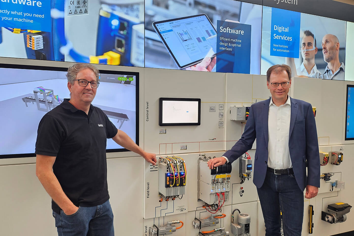 Zwei Männer vor einer technischen Ausstellung mit Hardware- und Softwarelösungen für Maschinen, im Hintergrund Bildschirme mit Informationen zu digitalen Diensten.