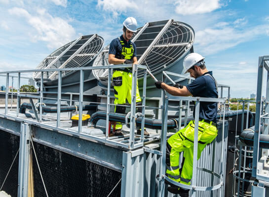 Two people wearing hard hats and high-visibility vests are standing on metal scaffolding and working on a ventilation system