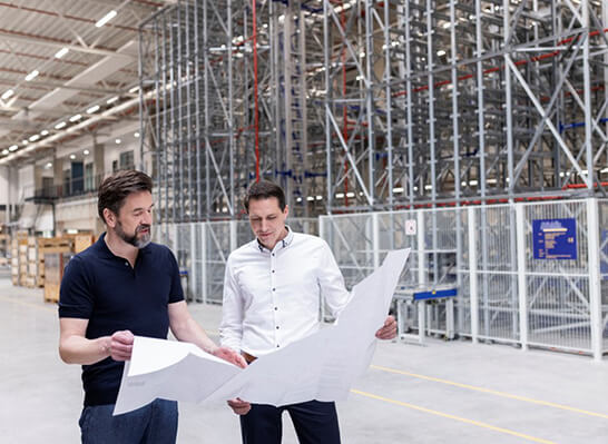 Two people in a warehouse looking at a blueprint for a modular machine.