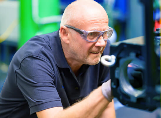 Mann mit Schutzbrille untersucht konzentriert ein mechanisches Bauteil in einer Werkstatt