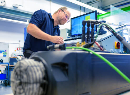 Mann mit Brille in Arbeitskleidung steht an einer Maschine mit Monitor und mehreren Kabeln