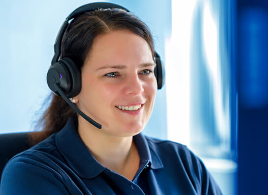 Frau mit Headset l&auml;chelt freundlich in die Kamera, w&auml;hrend sie an einem Computerbildschirm im B&uuml;ro arbeitet.