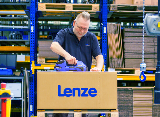 Mann verpackt sorgf&auml;ltig einen Karton mit der Aufschrift &bdquo;Lenze" in einem Lager mit Regalen voller Kartons.