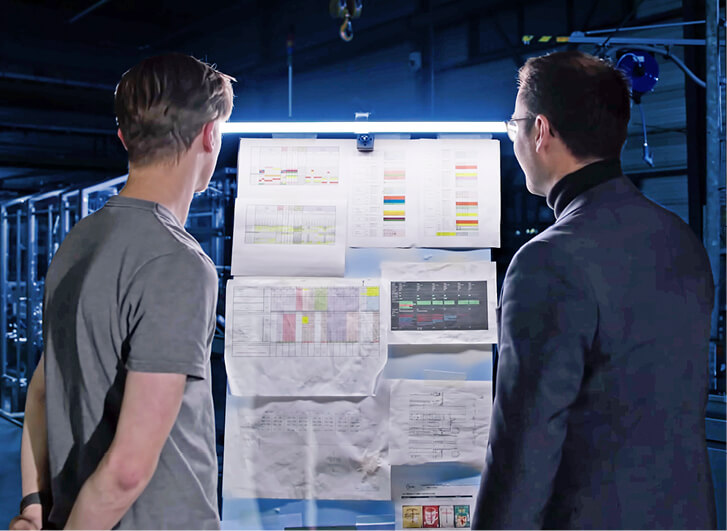 Two colleagues discussing technical drawings and diagrams in front of a screen.