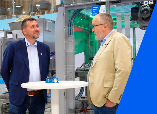 Two men are standing at a high-top table in front of a packaging machine and talking at a trade show.