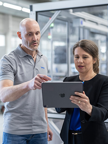 Zwei Personen vor einem industriellem Hintergrund, eine hält ein Tablet in der Hand, die andere Person zeigt mit dem Zeigefinger auf das Tablet